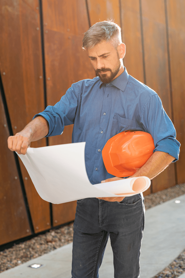 Mężczyzna w niebieskiej koszuli i kaskiem pod pachą czyta coś z dużego arkuszu papieru